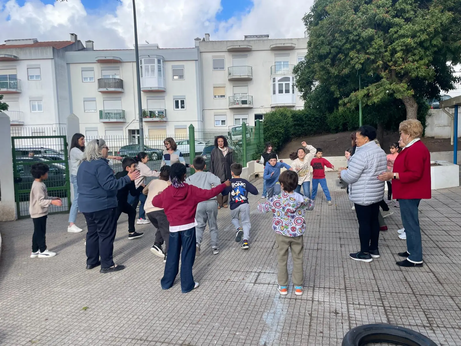 Des seniors et des enfants font une ronde en plein air, bras ouverts, dans la cour de récréation.